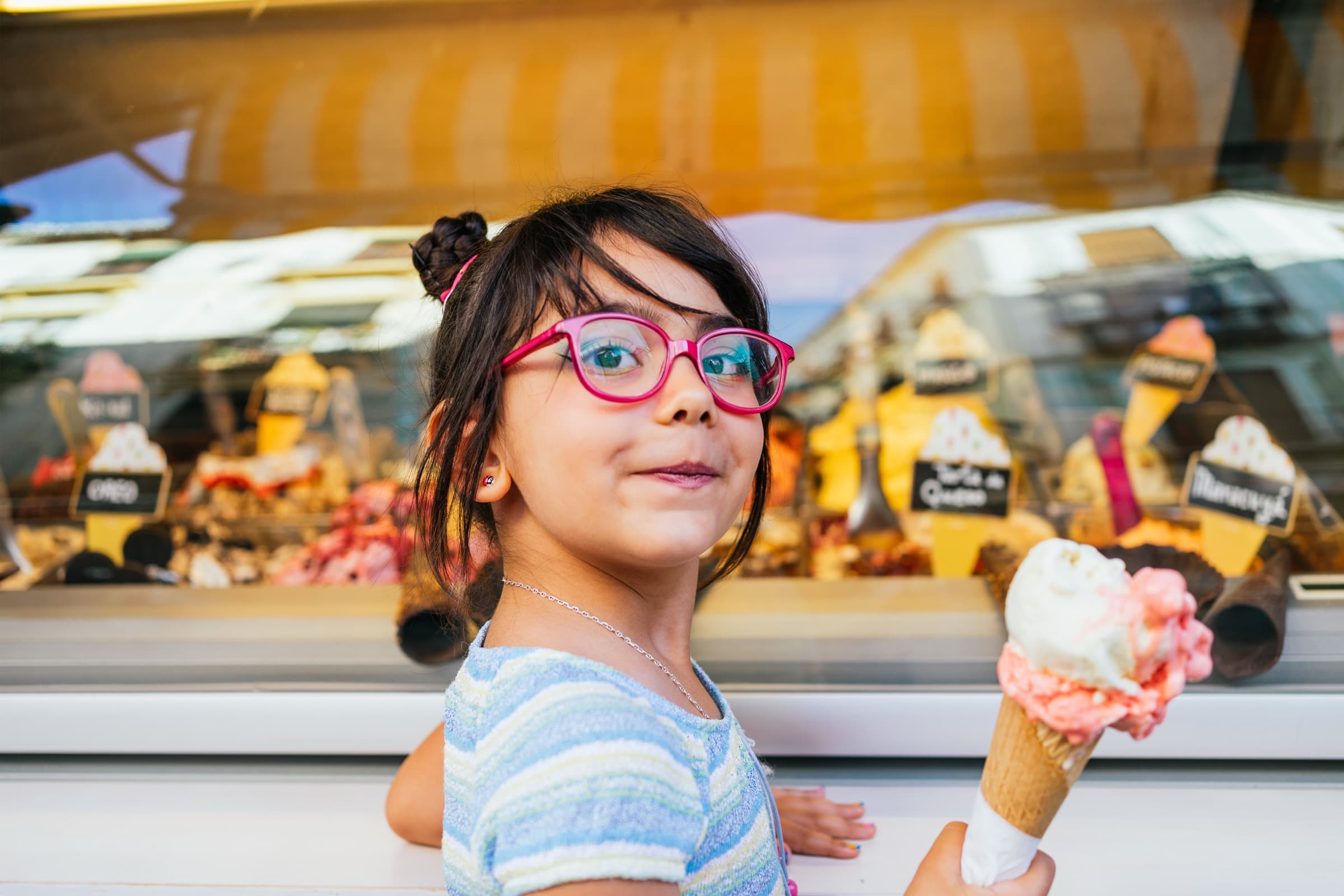 Une enfant souriante tient un cornet de glace devant la vitrine réfrigérée d’un glacier, les bacs de parfums alignés derrière elle.